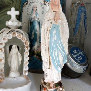 May include: A white porcelain figurine of the Virgin Mary, standing with hands clasped in prayer. She is wearing a blue robe with a white veil and is standing on a base with a floral design. The figurine is in front of a blue and white tin box with the words "Jesus, I am your Immaculate Conception" printed on it.