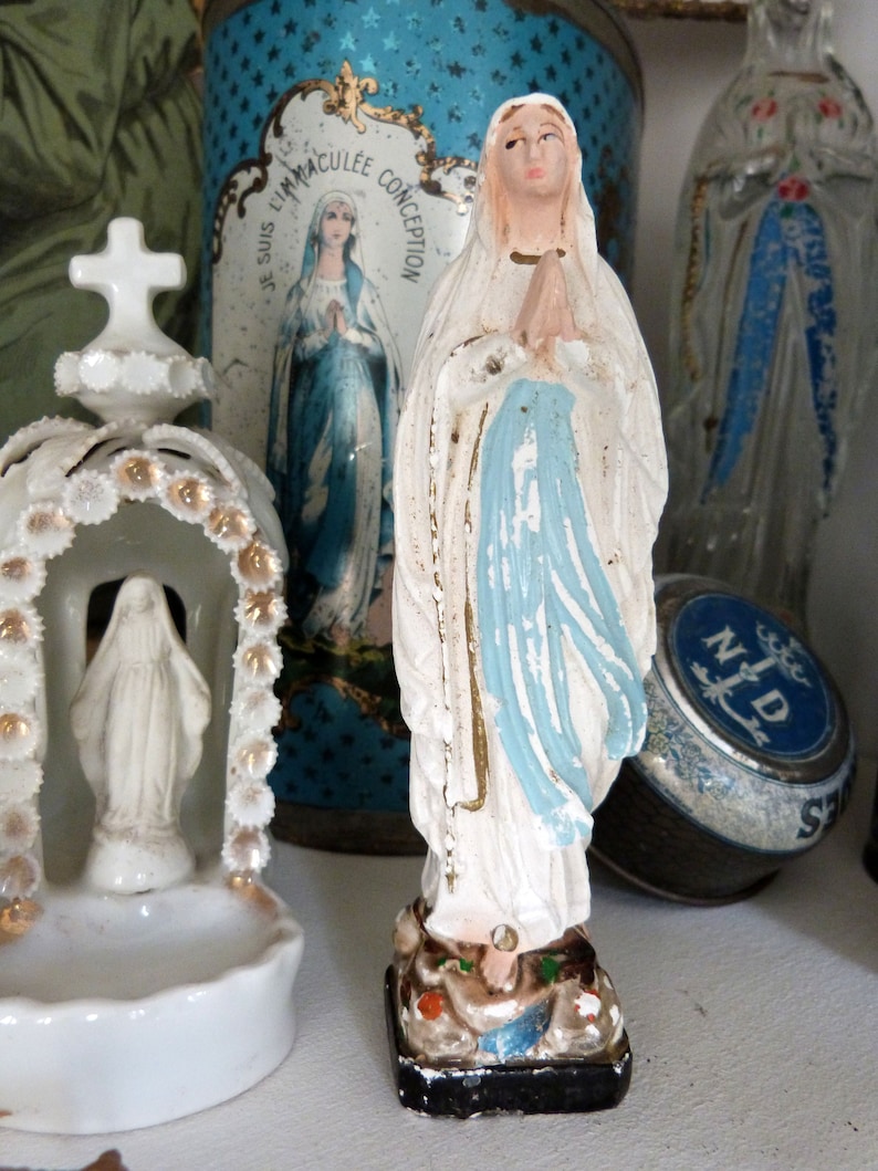 May include: A white porcelain figurine of the Virgin Mary, standing with hands clasped in prayer. She is wearing a blue robe with a white veil and is standing on a base with a floral design. The figurine is in front of a blue and white tin box with the words "Jesus, I am your Immaculate Conception" printed on it.