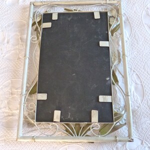May include: White metal picture frame with a decorative floral design. The frame has a black backing and is ready to hold a picture.