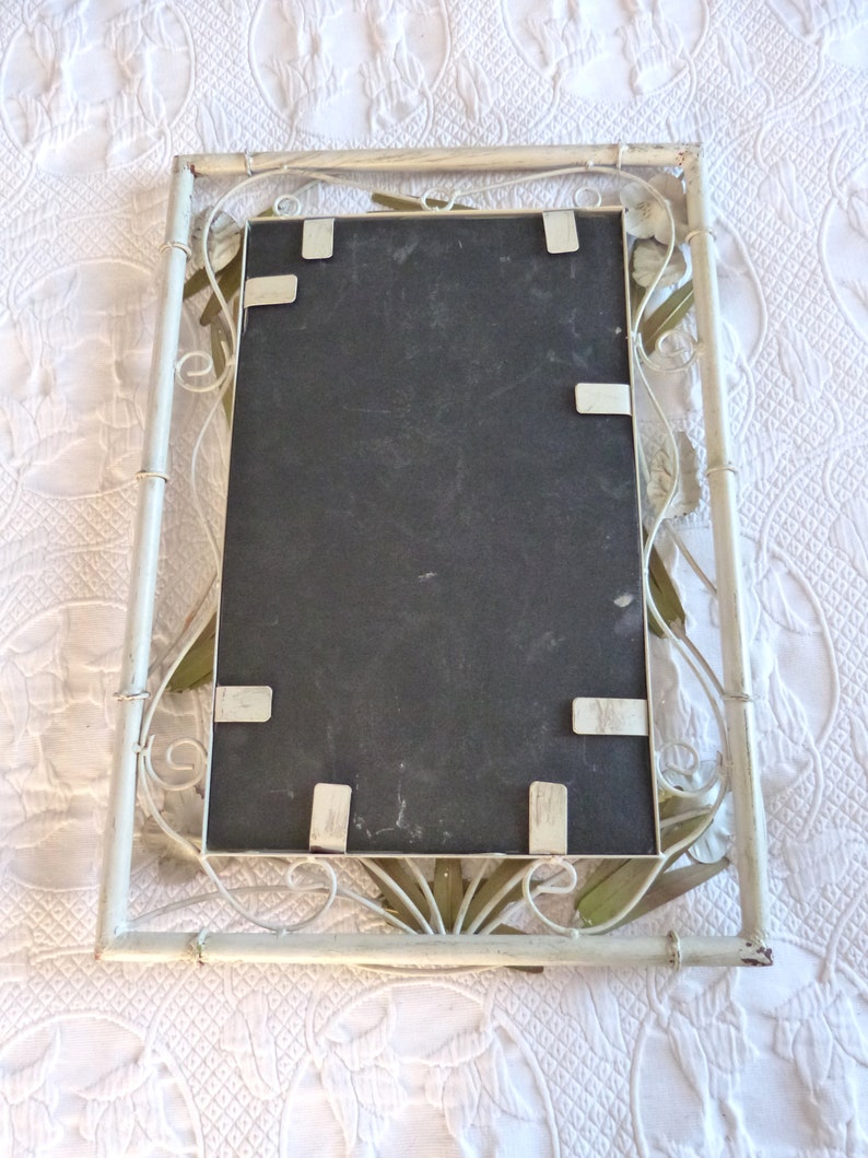 May include: White metal picture frame with a decorative floral design. The frame has a black backing and is ready to hold a picture.