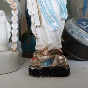 May include: White porcelain figurine of the Virgin Mary, wearing a blue robe and standing on a base with brown and green accents. The figurine is a religious icon.