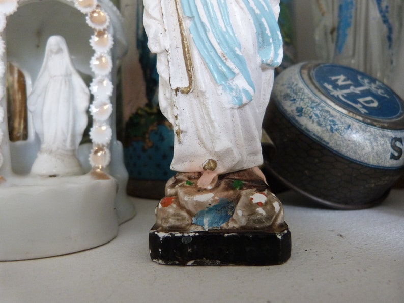 May include: White porcelain figurine of the Virgin Mary, wearing a blue robe and standing on a base with brown and green accents. The figurine is a religious icon.