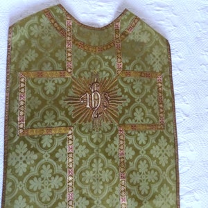 May include: Green liturgical vestment with a floral pattern and gold trim. The vestment features an embroidered gold IHS symbol at the center, surrounded by a sunburst design. The trim includes pink accents.