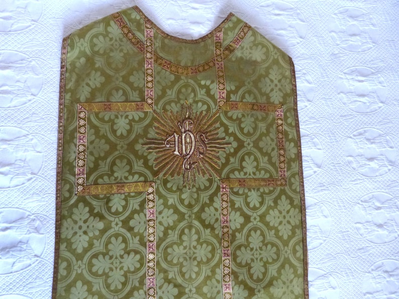 May include: Green liturgical vestment with a floral pattern and gold trim. The vestment features an embroidered gold IHS symbol at the center, surrounded by a sunburst design. The trim includes pink accents.