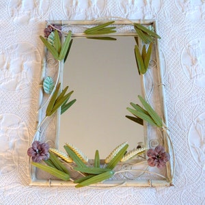 May include: A white metal framed mirror with a floral design. The frame is decorated with green and purple metal flowers and leaves.