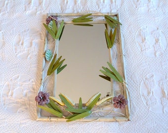 May include: A white metal framed mirror with a floral design. The frame is decorated with green and purple metal flowers and leaves.