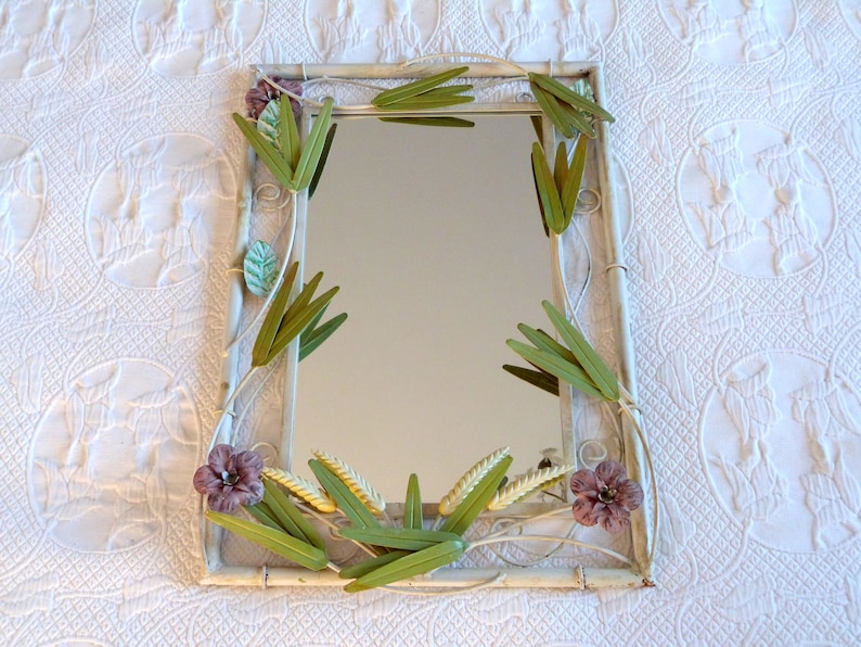 May include: A white metal framed mirror with a floral design. The frame is decorated with green and purple metal flowers and leaves.