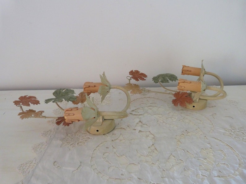 May include: Two vintage wall sconces with a leafy vine design. The sconces are painted in a light green and brown color scheme. Each sconce has two light bulbs.