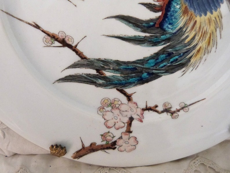 May include: A white porcelain plate with a blue and yellow bird perched on a branch with pink blossoms. The bird's tail feathers are spread out and the branch has thorns.