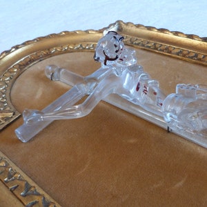 May include: A glass crucifix with a clear body and red accents. The crucifix is mounted in a gold-colored frame.
