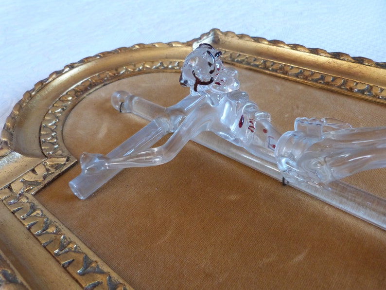 May include: A glass crucifix with a clear body and red accents. The crucifix is mounted in a gold-colored frame.