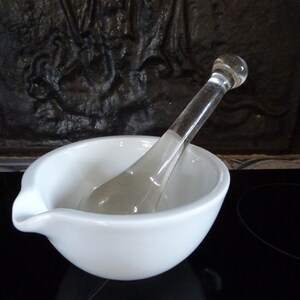 May include: A white ceramic mortar and pestle set with a clear glass pestle. The mortar has a spout for pouring.