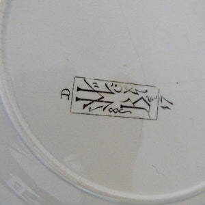 Pode incluir: Close-up da parte inferior de um prato de cer&acirc;mica branco com uma marca retangular marrom que diz "D" e um design estilizado.