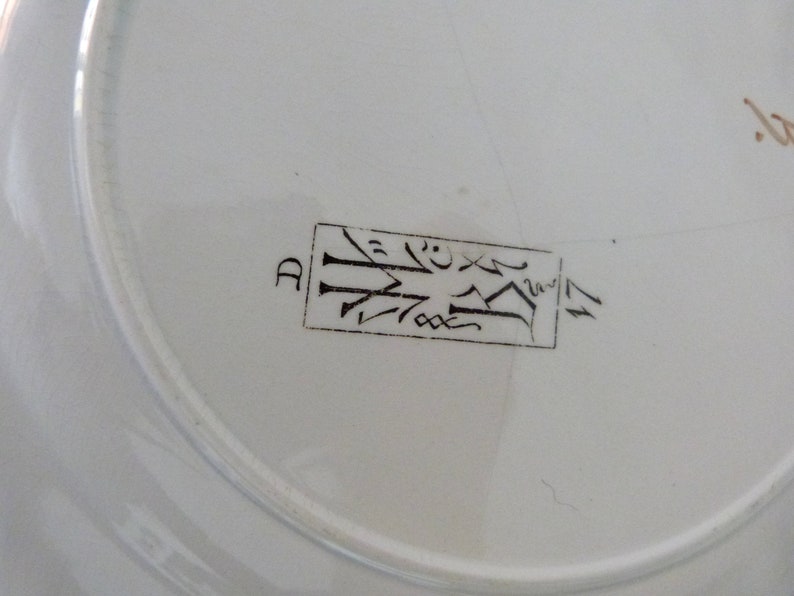 Pode incluir: Close-up da parte inferior de um prato de cer&acirc;mica branco com uma marca retangular marrom que diz "D" e um design estilizado.