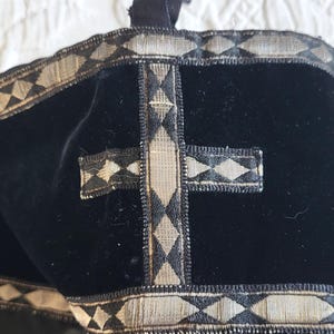 May include: Black velvet religious head covering with silver and black trim. The trim features a repeating diamond pattern. A silver cross is centered on the front. The head covering is likely used in religious ceremonies.