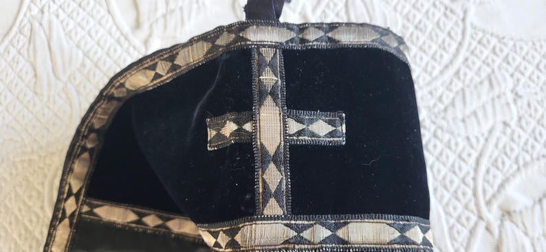 May include: Black velvet religious head covering with silver and black trim. The trim features a repeating diamond pattern. A silver cross is centered on the front. The head covering is likely used in religious ceremonies.