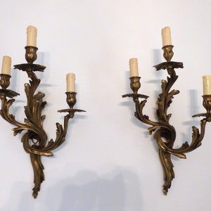 K&ouml;nnte beinhalten: Ein Paar verzierte goldene Wandleuchter mit jeweils drei Kerzenhaltern. Die Leuchter sind aus Metall und haben ein geschwungenes Design.