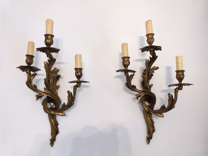 K&ouml;nnte beinhalten: Ein Paar verzierte goldene Wandleuchter mit jeweils drei Kerzenhaltern. Die Leuchter sind aus Metall und haben ein geschwungenes Design.