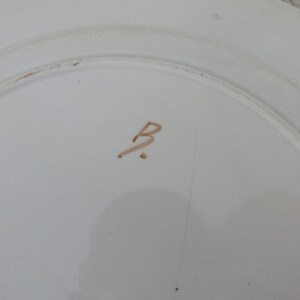 Pode incluir: Close-up da parte inferior de um prato de cer&acirc;mica branco com uma letra "B" escrita &agrave; m&atilde;o em marrom no centro.