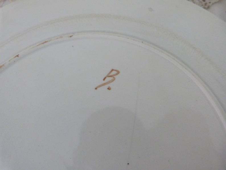 Pode incluir: Close-up da parte inferior de um prato de cer&acirc;mica branco com uma letra "B" escrita &agrave; m&atilde;o em marrom no centro.