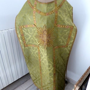 May include: A green liturgical vestment with gold and red embroidered details. The vestment features a floral pattern and a central embroidered emblem. The garment is displayed on a mannequin.