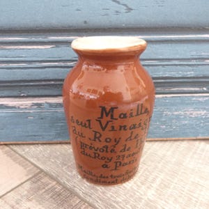 Puede incluir: Un tarro de cerámica marrón con borde blanco y texto negro. El tarro tiene un acabado brillante y una forma redondeada. El texto en el tarro dice "Maille, seul Vinaigre du Roy de..." y es un tarro de condimentos vintage.