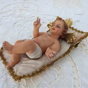 May include: A detailed figurine of a baby, lying on a cream-colored pillow with gold trim. The baby has light skin, a serene expression, and is wearing a white diaper. The pillow is set on a white, textured surface.