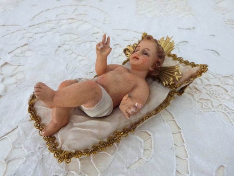 May include: A detailed figurine of a baby, lying on a cream-colored pillow with gold trim. The baby has light skin, a serene expression, and is wearing a white diaper. The pillow is set on a white, textured surface.