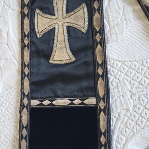 May include: Black and gold religious stole with a large cross emblem. The stole features a gold cross design on a black satin background, with gold trim and tassels. The stole is approximately 60 inches long.