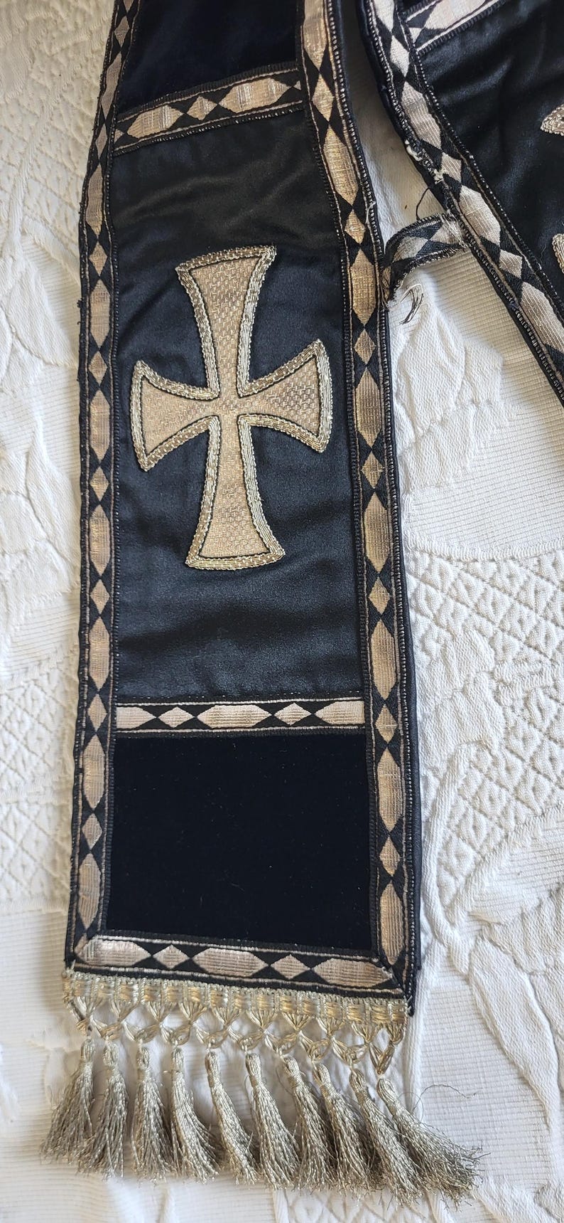 May include: Black and gold religious stole with a large cross emblem. The stole features a gold cross design on a black satin background, with gold trim and tassels. The stole is approximately 60 inches long.