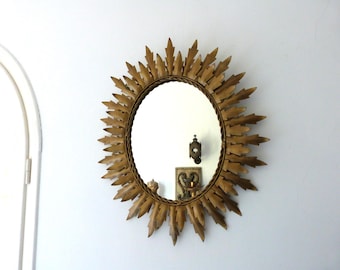 May include: A gold-toned sunburst mirror with an oval mirror center. The frame features a detailed design of stylized leaves radiating outwards. The mirror is mounted on a white wall, reflecting the room.