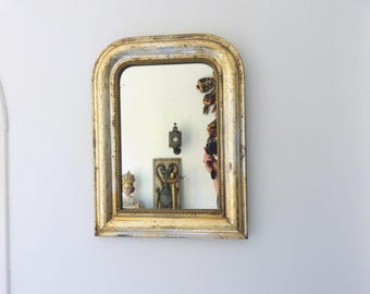 May include: An antique, arched-top mirror with a distressed gold-painted frame. The mirror reflects a room with decorative objects, including a small statue and wall sconces. The frame has a weathered appearance, adding to its vintage charm.