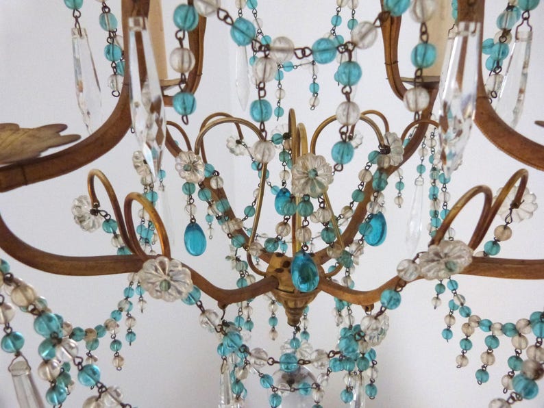 May include: Close-up of a vintage chandelier with a gold-toned metal frame. The chandelier is adorned with clear and turquoise-colored beads and crystal teardrops. The design includes floral accents and curved arms.