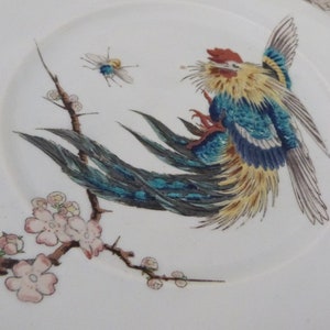 May include: A white ceramic plate with a hand-painted design of a blue and yellow bird with a long tail, perched on a branch with pink blossoms. A small blue and yellow insect is also painted on the plate.