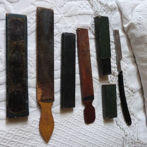 May include: A collection of seven vintage straight razor honing tools. The tools are made of wood and metal and have a variety of colours and patterns. The tools are arranged on a white surface.
