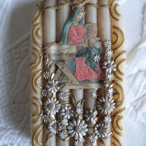 May include: A white candle with a raised design of a nativity scene with Mary, Joseph, and baby Jesus. The candle is decorated with gold and silver flowers.