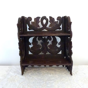 May include: A dark brown wooden shelf with intricate carved leaf designs. The shelf has two tiers and is perfect for displaying small items.