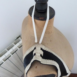 Może przedstawiać: W stylu vintage czarny i biały top typu halter z kremowym wykończeniem, eksponowany na manekinie. Top ma dekoracyjny wz&oacute;r i biały elastyczny pasek, kt&oacute;ry przechodzi wok&oacute;ł szyi. Manekin jest w kolorze jasnobrązowym.