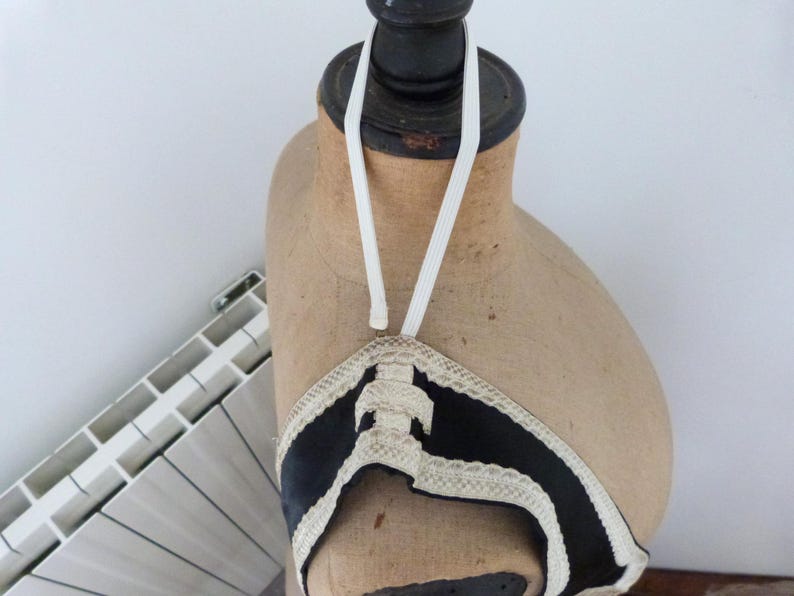 Może przedstawiać: W stylu vintage czarny i biały top typu halter z kremowym wykończeniem, eksponowany na manekinie. Top ma dekoracyjny wz&oacute;r i biały elastyczny pasek, kt&oacute;ry przechodzi wok&oacute;ł szyi. Manekin jest w kolorze jasnobrązowym.