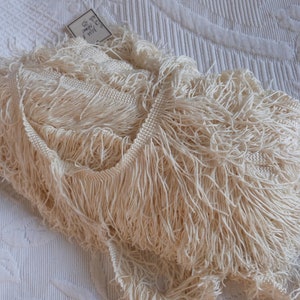 May include: A close-up of a pile of ivory white fringe trim. The trim is made of soft, delicate fibers and has a slightly textured appearance. The trim is perfect for adding a touch of elegance to any project.