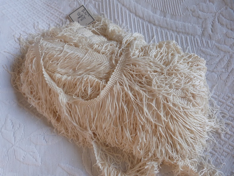 May include: A close-up of a pile of ivory white fringe trim. The trim is made of soft, delicate fibers and has a slightly textured appearance. The trim is perfect for adding a touch of elegance to any project.