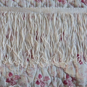 May include: White fringe trim with a subtle sheen. The fringe is made of small, tightly woven strands of fabric.