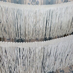May include: White fringe trim with a woven texture. The fringe is made of thin, white threads and is approximately 1 inch in length.