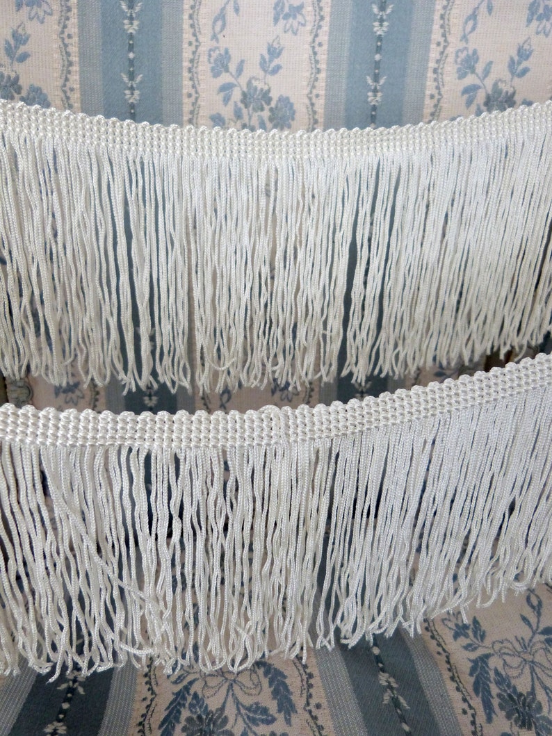 May include: White fringe trim with a woven texture. The fringe is made of thin, white threads and is approximately 1 inch in length.