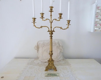 May include: A tall, gold-toned metal candelabra with five arms and a decorative base. The candelabra is designed to hold candles.