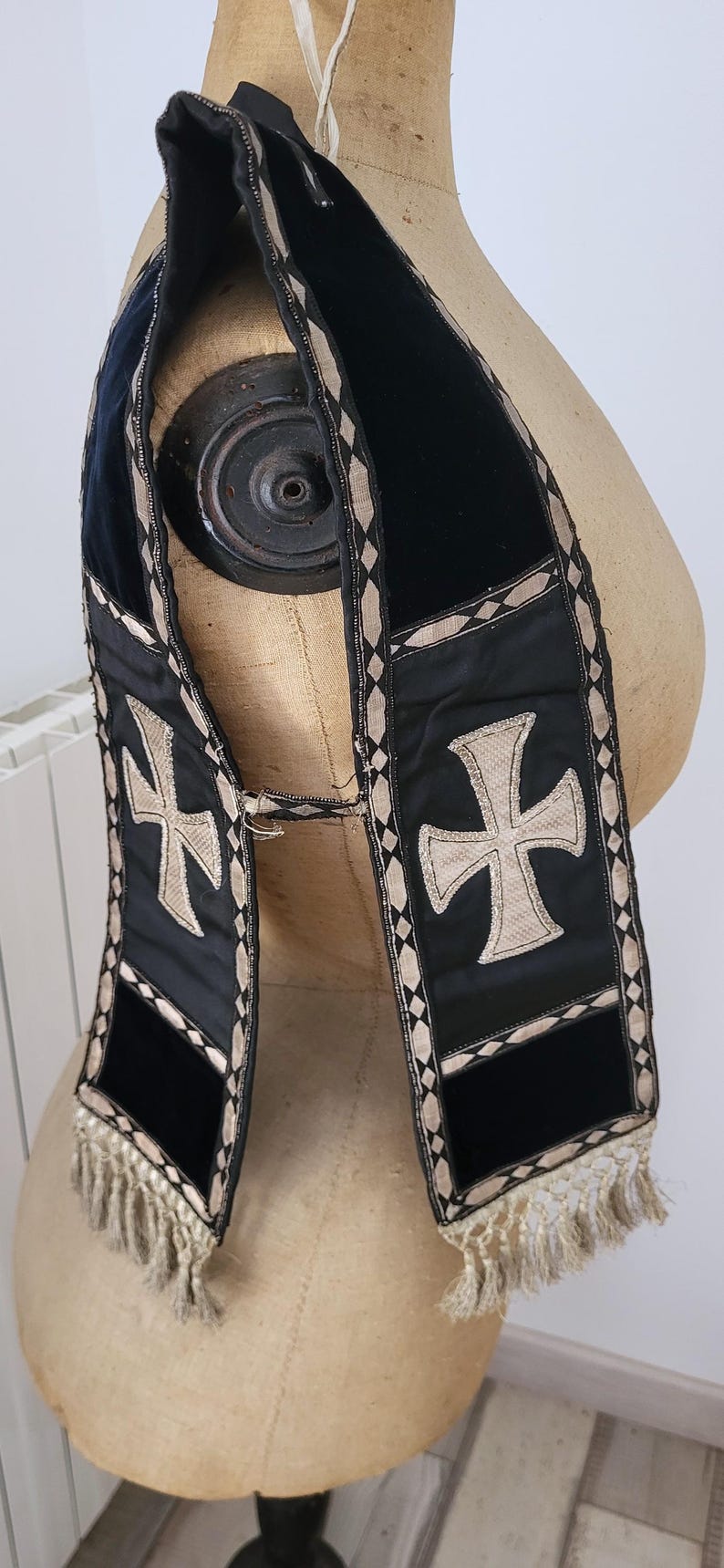 May include: Black velvet stole with a gold cross design and diamond-patterned trim. The stole has a fringe at the bottom and is displayed on a mannequin. A religious garment.
