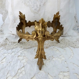 Op de afbeelding: Omsiers gouden metalen decoratieve standaard met een gedetailleerd gezicht en bladeren. De standaard heeft een cirkelvormige bovenkant en drie poten. De standaard is waarschijnlijk voor het tentoonstellen van een klein object.