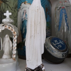 May include: White porcelain figurine of the Virgin Mary, wearing a white robe and standing on a black base. The figurine is in front of a blue and white tin with the words "Immacul&eacute;e Conception" on it.