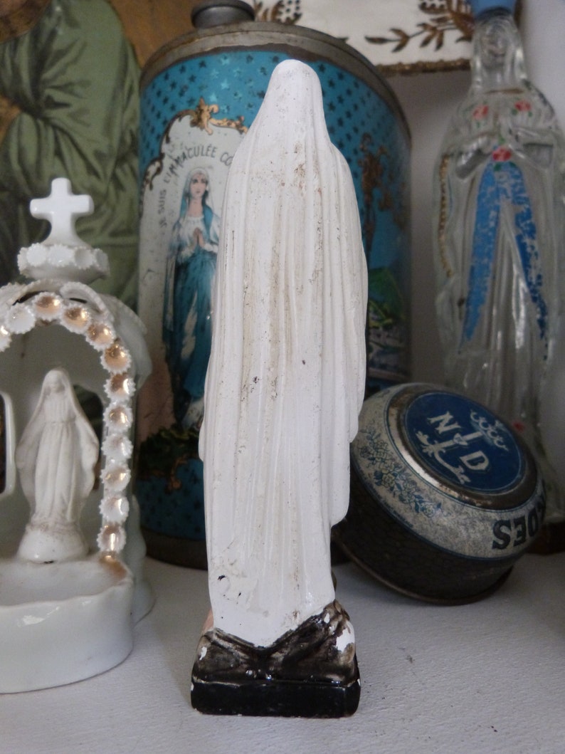 May include: White porcelain figurine of the Virgin Mary, wearing a white robe and standing on a black base. The figurine is in front of a blue and white tin with the words "Immacul&eacute;e Conception" on it.