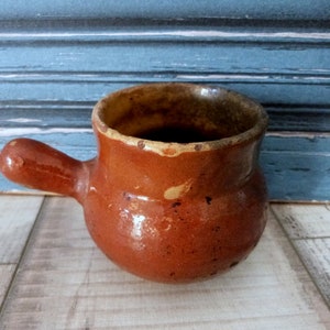 May include: A small, brown earthenware pot with a single handle. The pot has a rustic, handmade look and is glazed in a brown and yellow color.